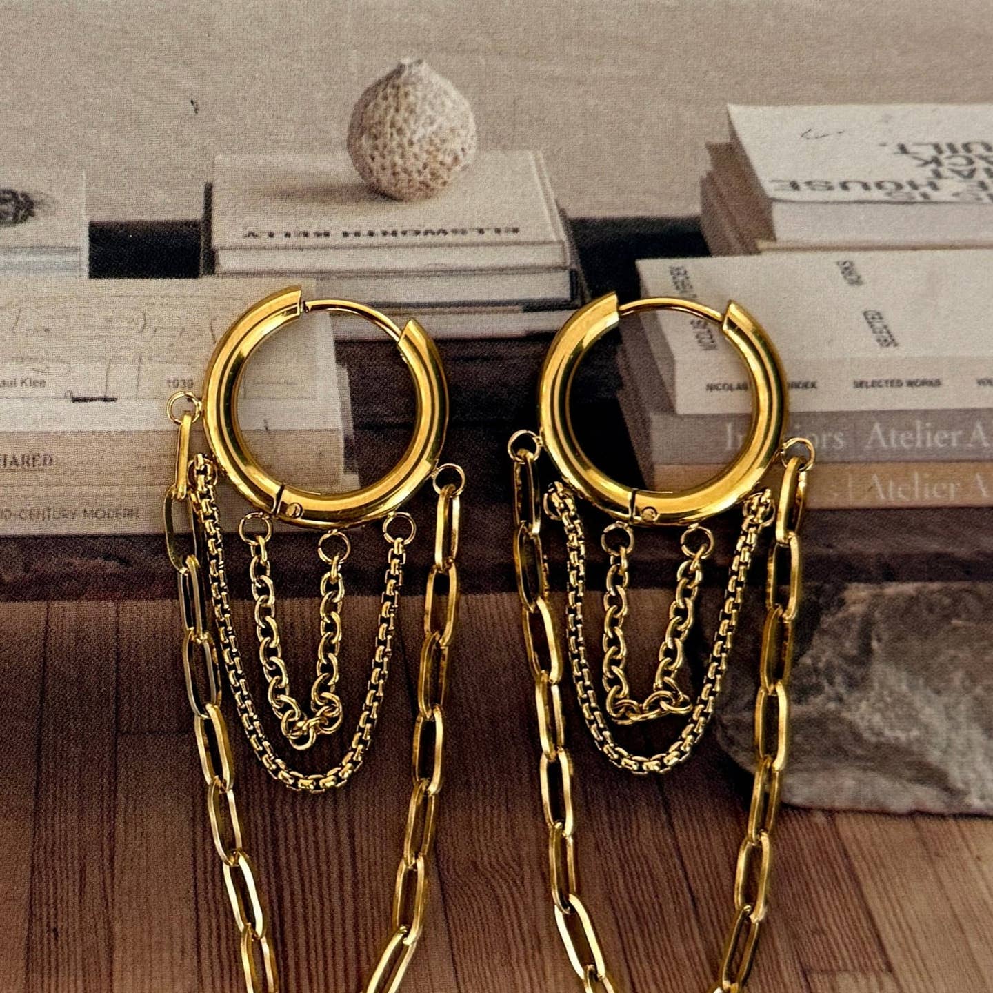 Gold hoop earrings with chains on a wooden surface with books in the background