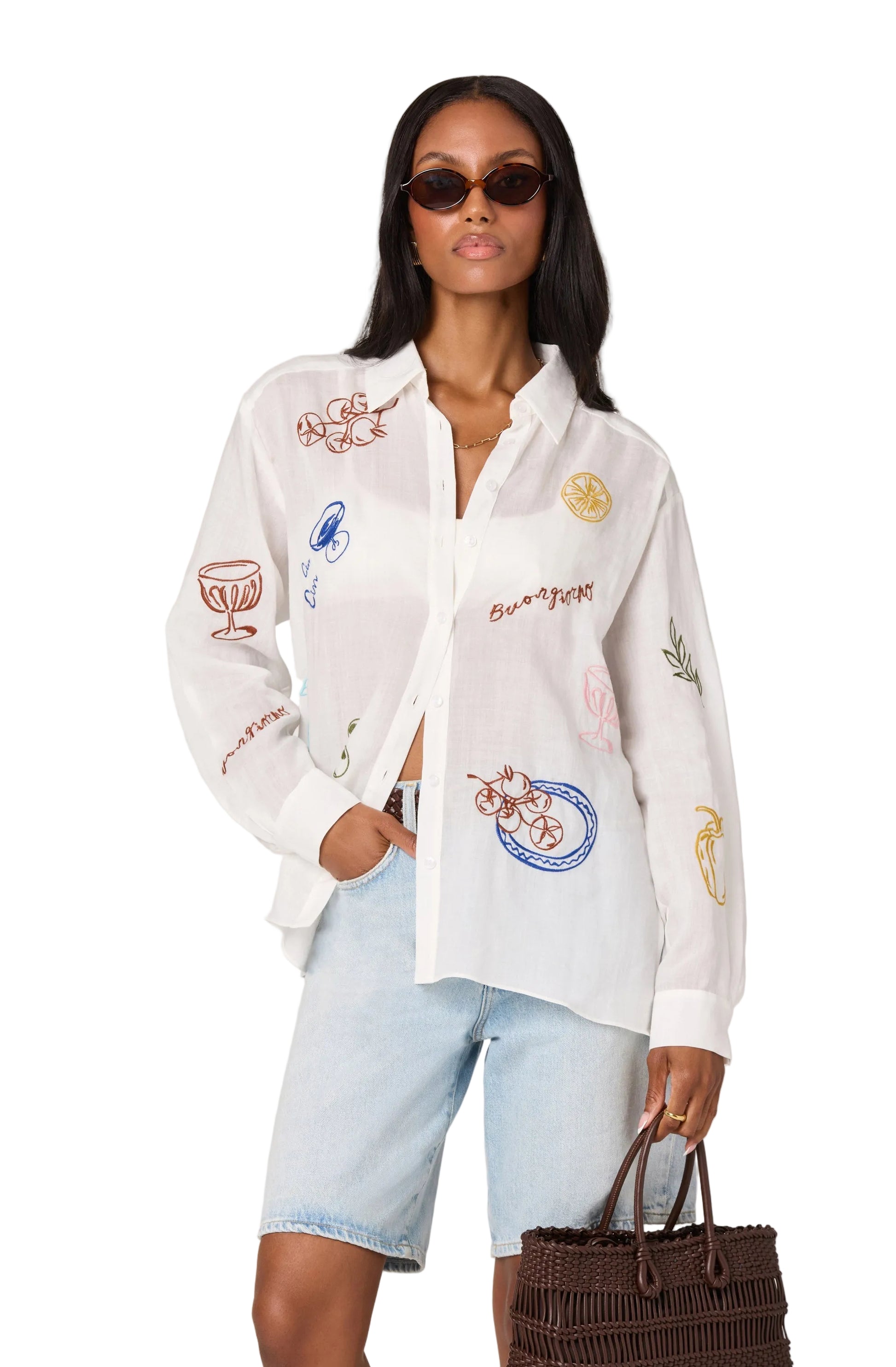 Woman wearing a white embroidered shirt with light blue shorts and sunglasses, holding a brown bag on a white background.