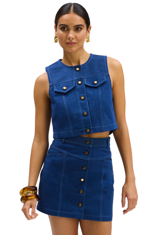 A model wearing a matching blue denim-like two-piece set consisting of a sleeveless cropped top with button details and a high-waisted mini skirt with front buttons, photographed against a plain white background.