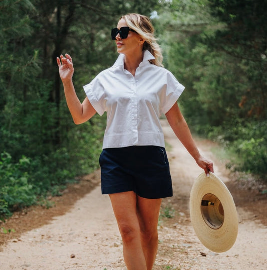 Woman in white shirt and black shorts walking on a dirt path with a hat in her hand.