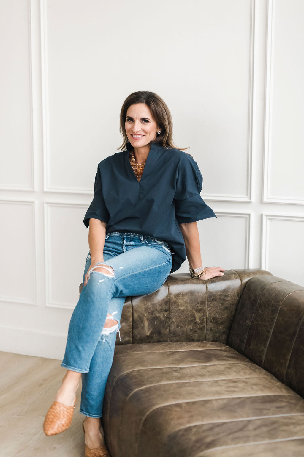 Woman sitting on a brown leather couch wearing a navy blue top and ripped jeans.