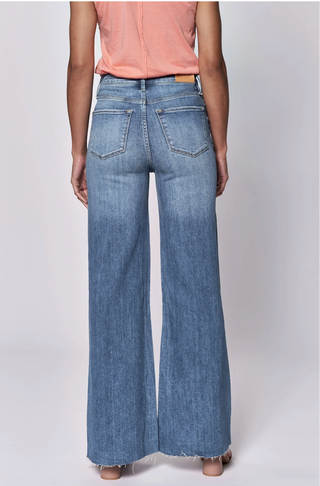 A person shown from the back wearing wide-leg blue jeans with a peach top, on a light gray studio background. Product text below reads 'FIONA WIDE LEG JEAN NATAL' with a review rating and price.