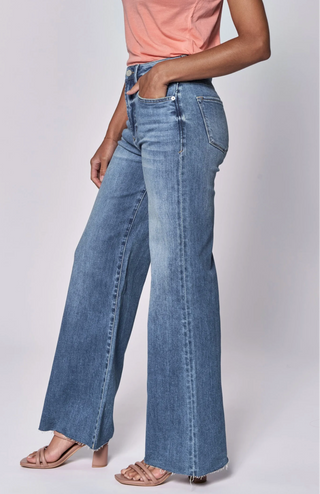A model wearing Fiona wide-leg jeans in a light blue wash, paired with a peach top and beige sandals, shown on a light gray background with a product info overlay at the bottom.